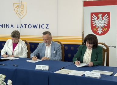 Trzy osoby siedzą przy niebieskim stole i podpisują dokumenty. W tle biało-żółto-granatowy baner z graficznym logotypem po lewej i czerwonym herbem z białym orłem po prawej. Wnętrze sali, widoczne krzesła i dokumenty na stole.