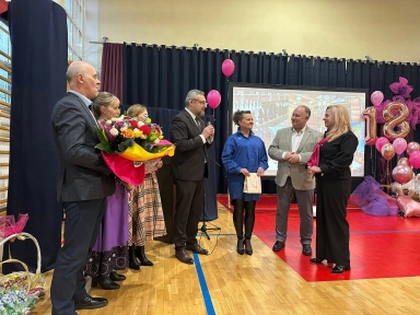 Siedem osób stoi w sali gimnastycznej podczas uroczystości. Trzy kobiety i czterech mężczyzn stoi przed ciemnymi zasłonami i ekranem projekcyjnym. Po prawej widoczne różowe balony i duża złota cyfra „18”; podłoga drewniana z czerwoną matą.