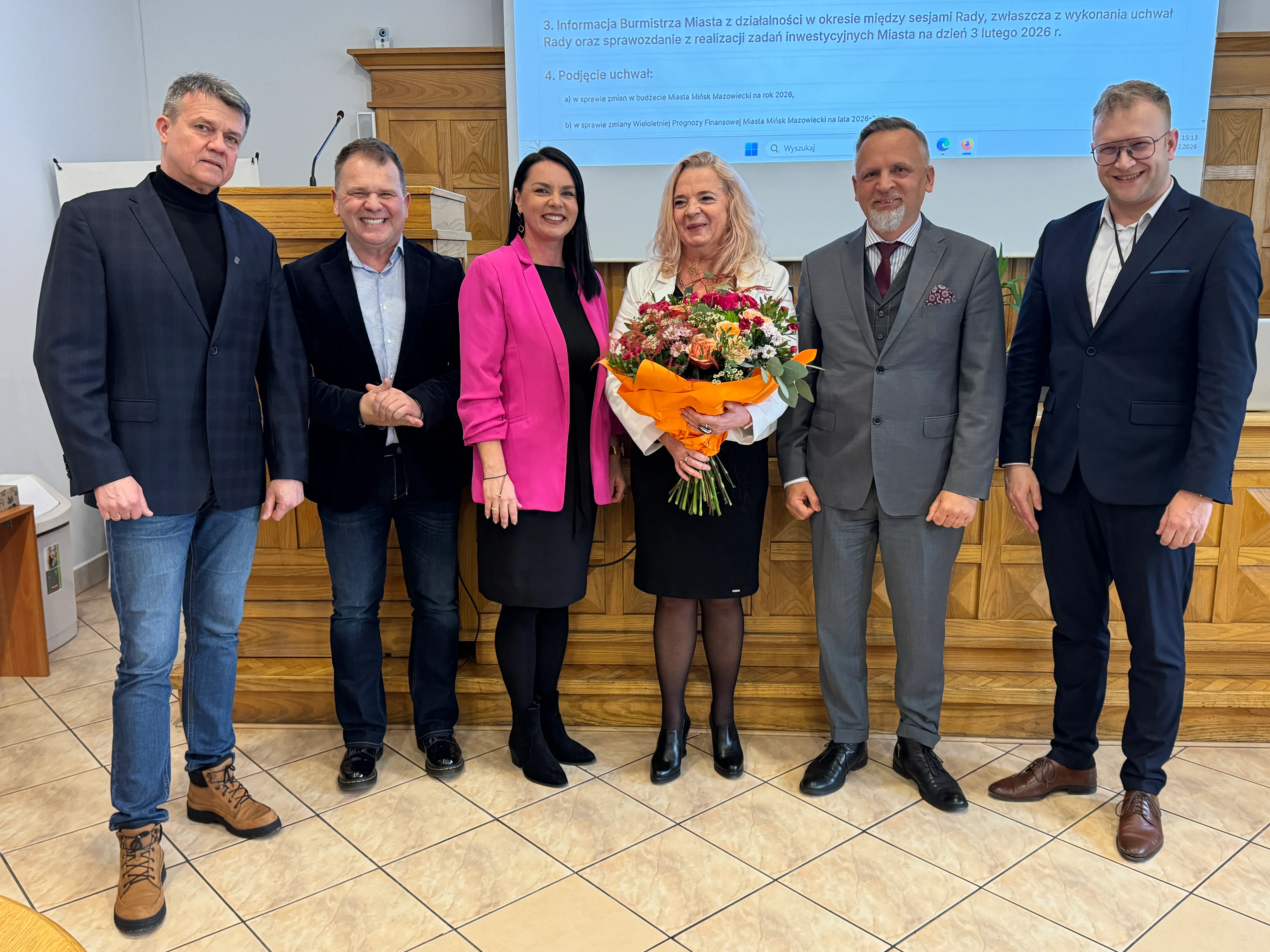 Sześć osób stoi w rzędzie w sali konferencyjnej. Kobieta w środku trzyma duży bukiet kwiatów owinięty w pomarańczowy papier. Kobieta obok po lewej nosi różową marynarkę, pozostali mężczyźni mają garnitury lub marynarki i eleganckie buty. W tle widać drewniany pulpit, ekran z niebieskim tekstem i jasną, kafelkową podłogę.