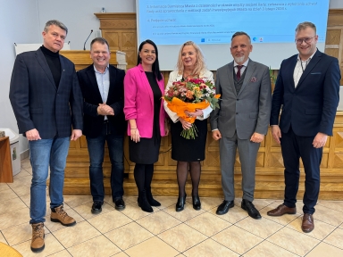 Sześć osób stoi w rzędzie w sali konferencyjnej. Kobieta w środku trzyma duży bukiet kwiatów owinięty w pomarańczowy papier. Kobieta obok po lewej nosi różową marynarkę, pozostali mężczyźni mają garnitury lub marynarki i eleganckie buty. W tle widać drewniany pulpit, ekran z niebieskim tekstem i jasną, kafelkową podłogę.