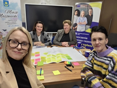 Cztery kobiety siedzą przy drewnianym stole w sali konferencyjnej. Kobieta z przodu po lewej robi selfie i ma okulary. Kobieta z przodu po prawej ma kolorowy pasiasty sweter. Dwie kobiety siedzą z tyłu przy stole. Na stole leży duży arkusz papieru z żółtymi, różowymi i zielonymi karteczkami samoprzylepnymi oraz flamastry i długopisy. W tle stoi duży czarny ekran i dwa stojące banery.