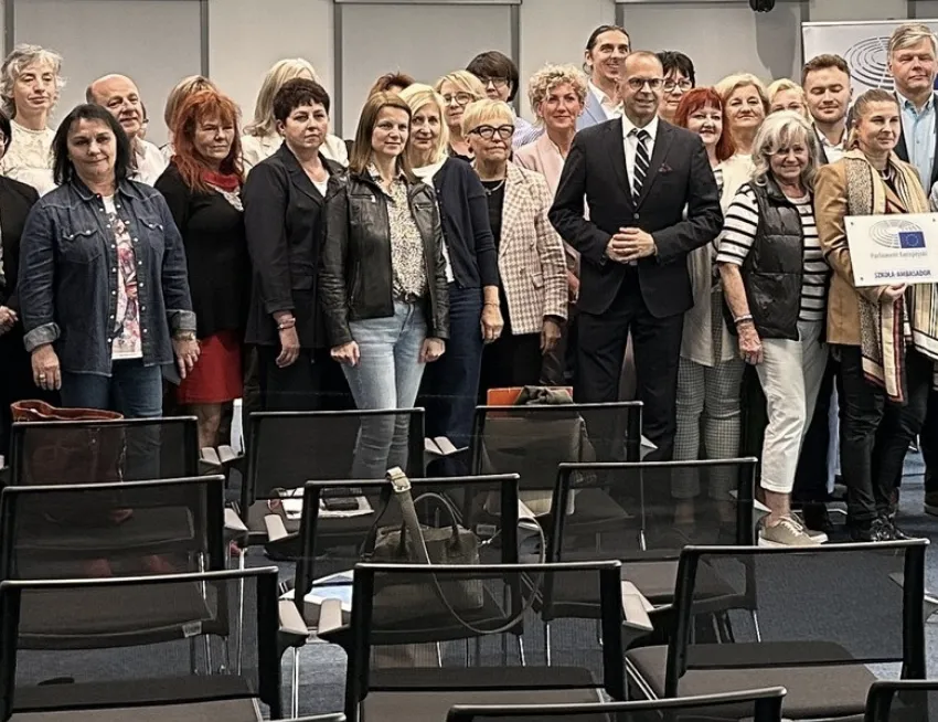 Grupa kilkunastu osób stojących w rzędzie za pustymi krzesłami w pomieszczeniu konferencyjnym.