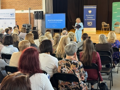 Publiczność siedzi na składanych krzesłach w sali z drewnianą sceną. Kobieta w jasnoniebieskim garniturze stoi na scenie i czyta z teczki. Obok stoi ekran z prezentacją i kilka banerów z herbem i logotypami. Widzowie to głównie dorośli, siedzą tyłem do aparatu.