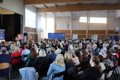 W sali gimnastycznej wiele osób siedzi w rzędach i słucha prelegentki przy mównicy. Jasne okna i drewniane belki sufitowe widoczne są nad aulą, z bocznymi banerami i koszem do koszykówki przy ścianie.