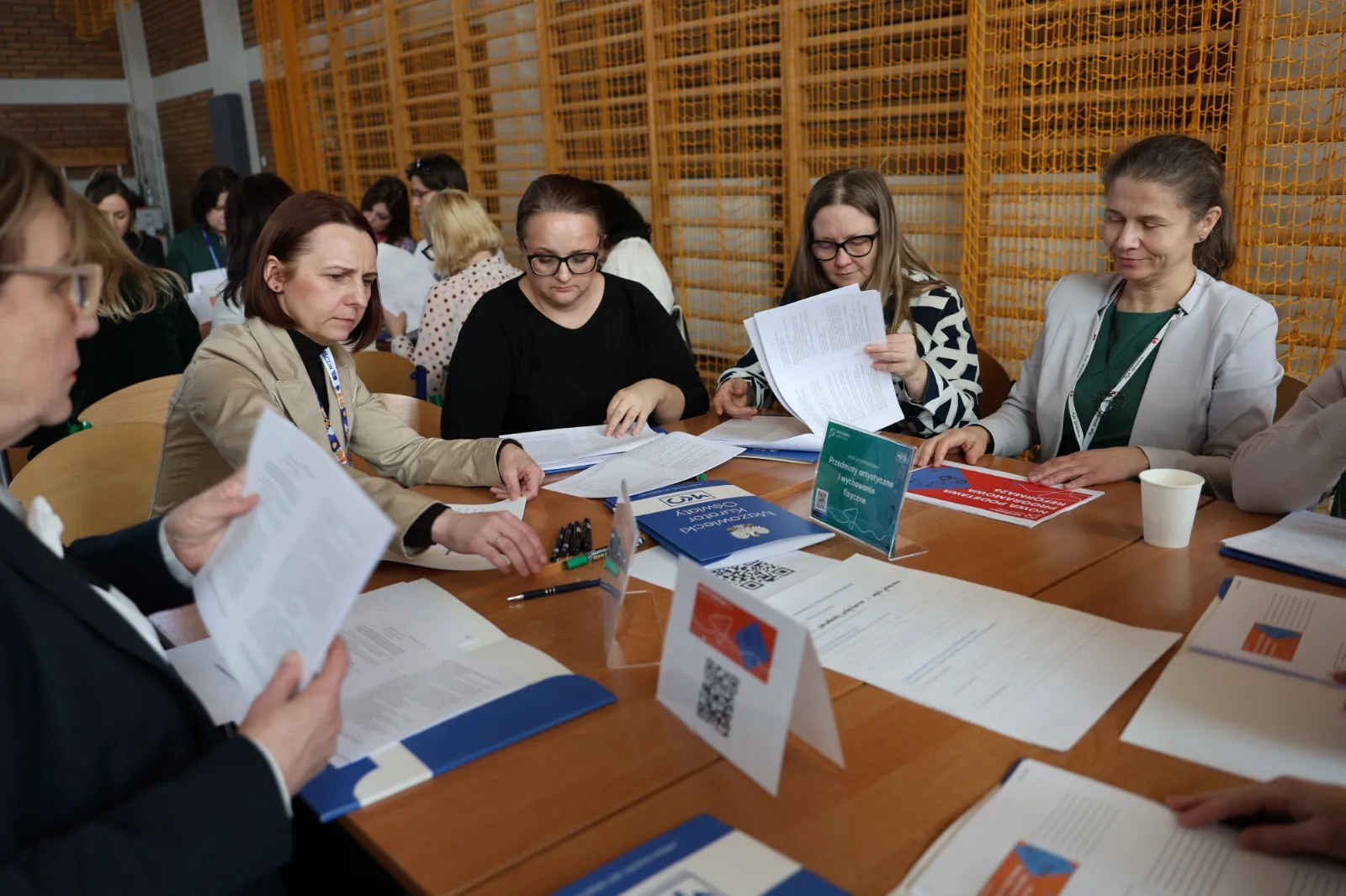 Pięć kobiet siedzi przy dużym drewnianym stole i przegląda dokumenty. W tle siatka i sprzęt sali gimnastycznej, na stole kartki, teczki, identyfikatory i kubek.