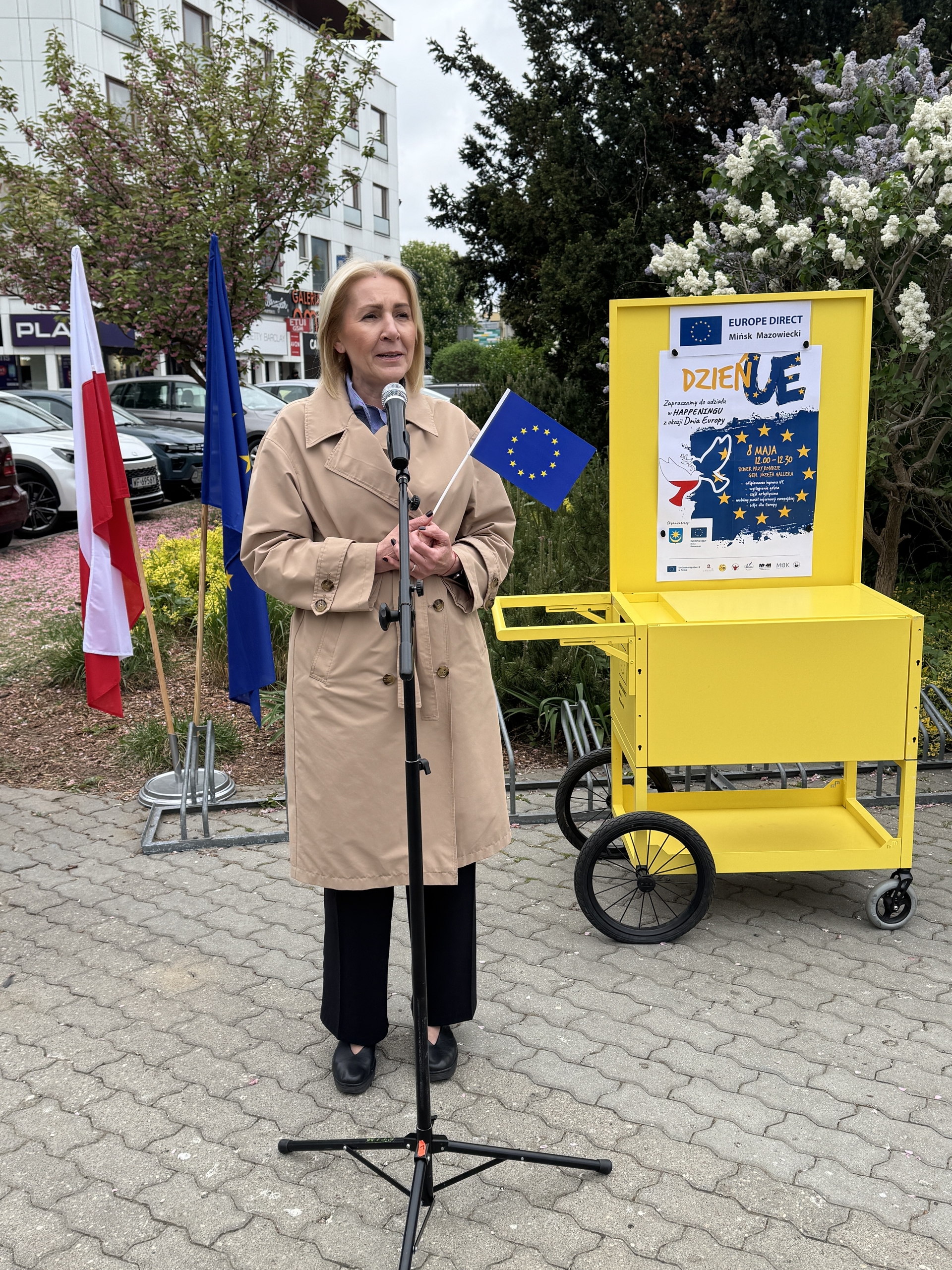 Kobieta stoi przy mikrofonie na zewnątrz w ciągu dnia. Ma beżowy płaszcz i trzyma małą flagę Unii Europejskiej. Obok stoją flagi Polski i UE, żóły wózek z plakatem i kwitnące krzewy w tle.