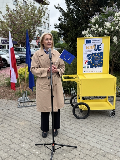 Kobieta stoi przy mikrofonie na zewnątrz w ciągu dnia. Ma beżowy płaszcz i trzyma małą flagę Unii Europejskiej. Obok stoją flagi Polski i UE, żóły wózek z plakatem i kwitnące krzewy w tle.