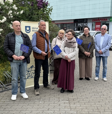 Sześć dorosłych osób stoi na zewnątrz przed krzewami i nowoczesnym budynkiem. Trzymają małe niebieskie flagi z żółtymi gwiazdami. Obok wisi materiałowy baner. Scena ma dzienną, miejską scenerię. Grupa stoi blisko siebie i wygląda poważnie.