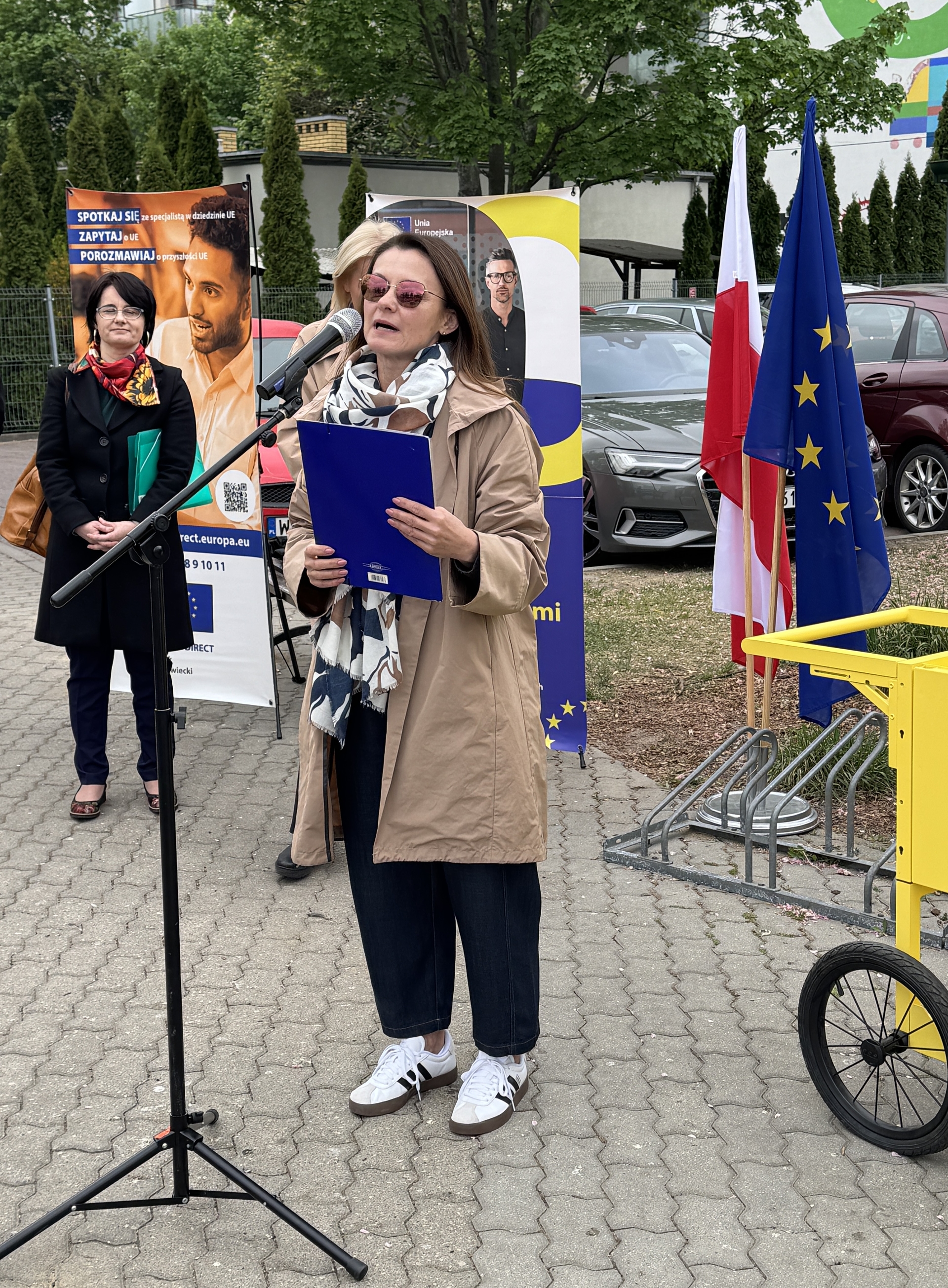 Kobieta w beżowym płaszczu i szaliku mówi do mikrofonu trzymając niebieski teczkę na zewnątrz. W tle stoją flagi UE i Polski, banery informacyjne, zaparkowane samochody oraz druga osoba obserwująca.