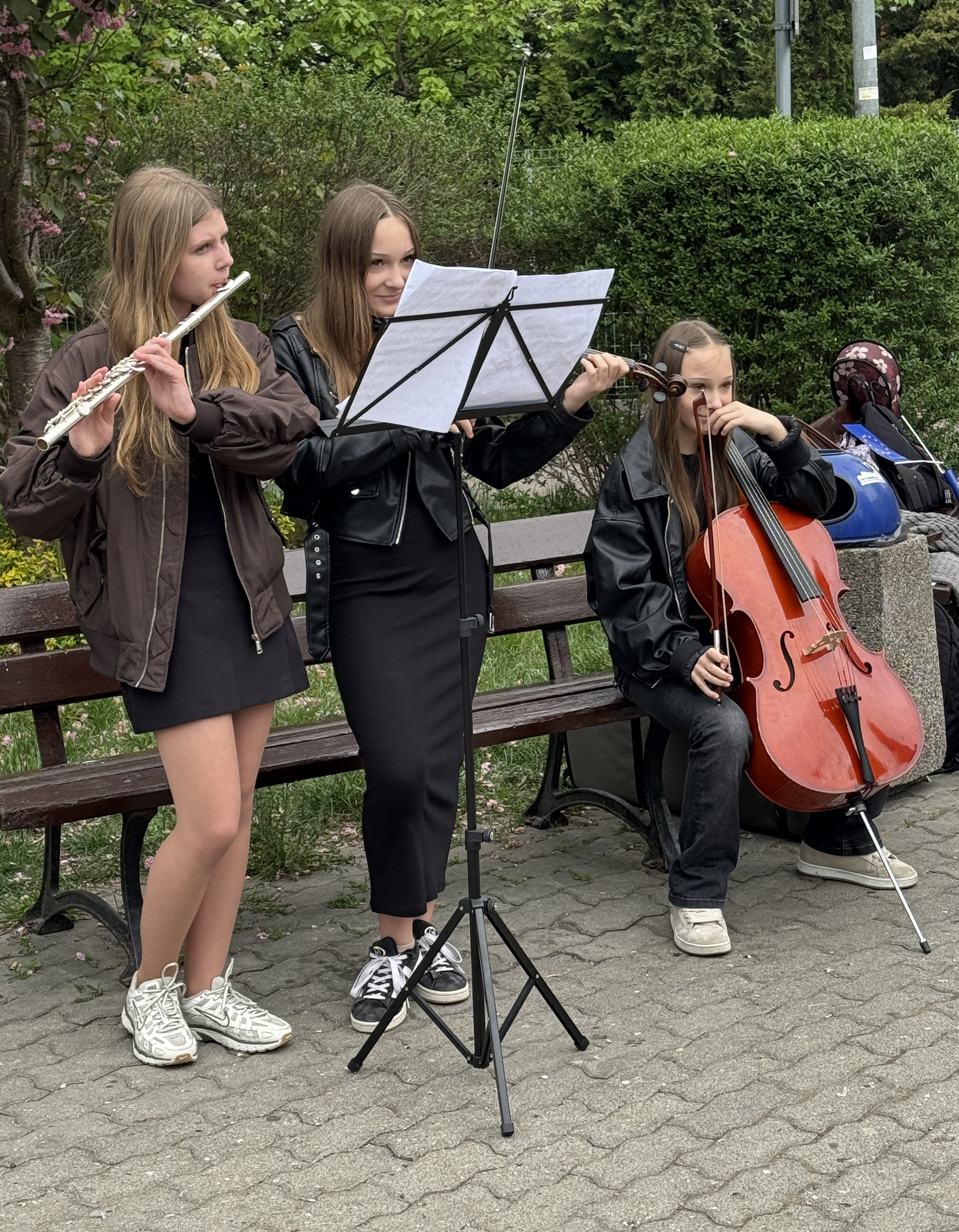 Trzy dziewczyny grają na instrumentach przy ławce w parku. Jedna stoi z fletem po lewej, jedna stoi ze skrzypcami i nutami na pulpicie na środku, trzecia siedzi z wiolonczelą po prawej. Tło stanowią zielone krzewy i drzewa, dzień, naturalne światło.