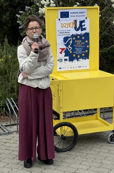 Kobieta w okularach trzyma mikrofon i przemawia. Stoi na zewnątrz przy żółtym pojeździe/kiosku z przytwierdzonym plakatem. W tle krzewy i drzewa, dzień.
