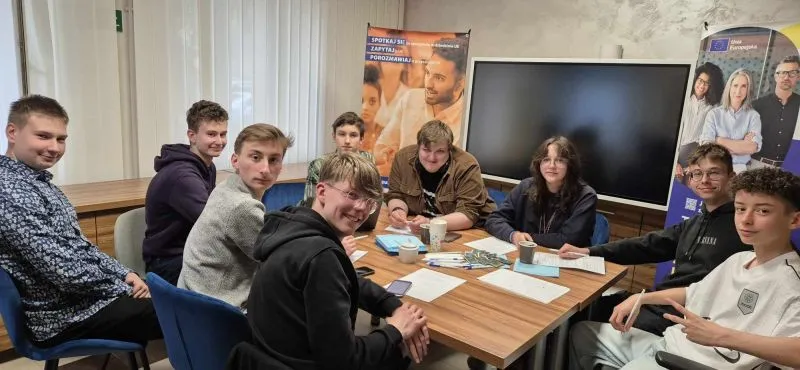 Dziewięć nastolatków siedzi przy stole z dokumentami i kubkami, prowadząc dyskusję.