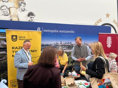 Pięć osób rozmawia przy stoisku wystawowym w hali. Za nimi duży niebieski baner z panoramą miasta i żółty baner z herbem. Na stole koszyki z przekąskami, chleb i ulotki. W górnej części ściany mural z czarno-białymi portretami sportowców.
