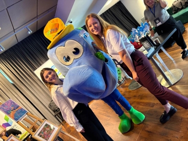 Dwie uśmiechnięte kobiety stoją po obu stronach postaci w dużym niebieskim kostiumie z dużymi oczami i żółtym kapeluszem, w pomieszczeniu z drewnianą podłogą i wystawionymi obrazami oraz stołem z dekoracjami.