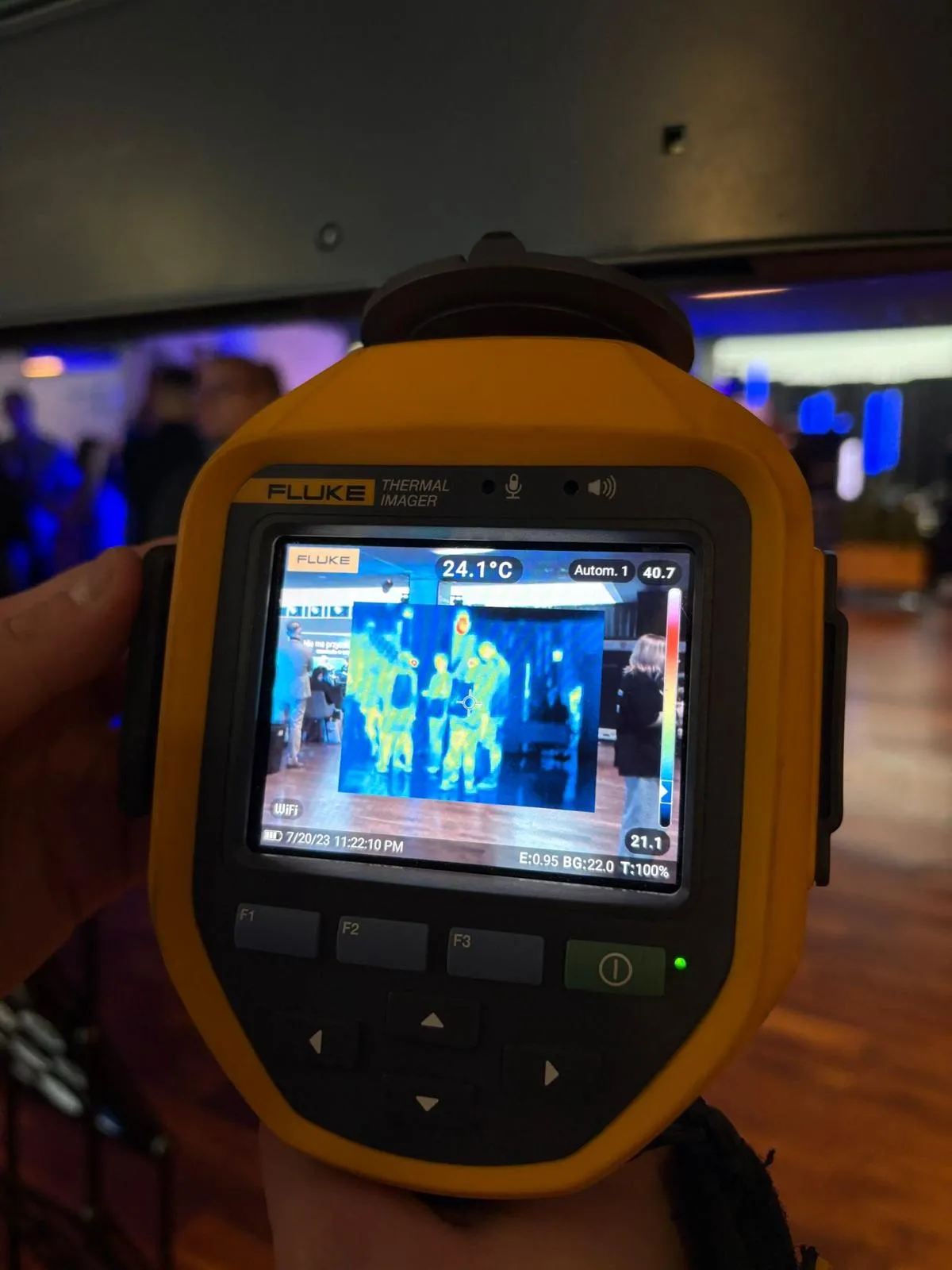 Ręka trzyma żółty miernik termowizyjny marki Fluke z wyświetlaczem pokazującym kolorowy obraz termiczny grupy osób w pomieszczeniu o ciemnym wnętrzu.
