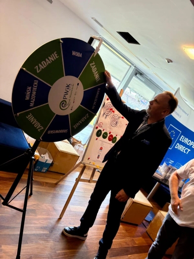 Mężczyzna w garniturze obraca koło fortuny ustawione na stojaku w jasnym pomieszczeniu z drewnianą podłogą. W tle tablica na stojaku i niebieski baner z napisem Europe Direct. Obok stoi druga osoba w białej koszulce.