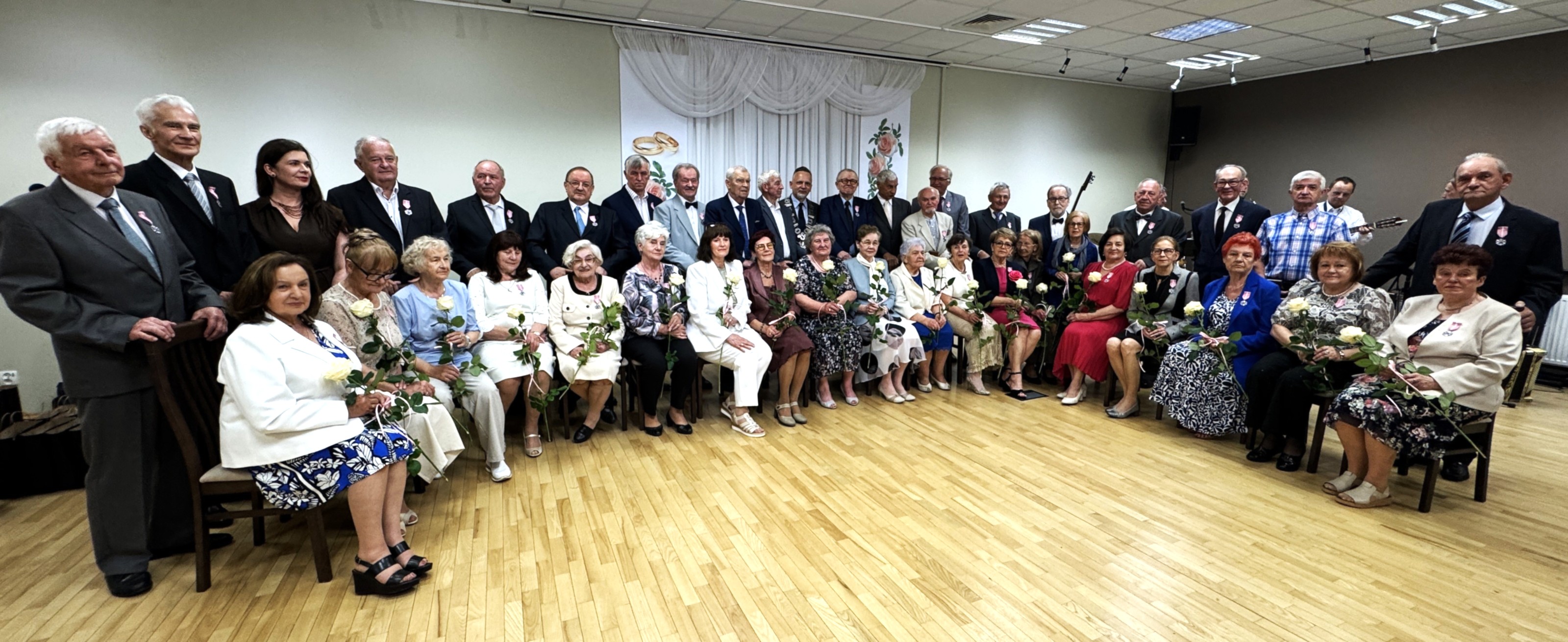 Starsze kobiety i mężczyźni siedzą i stoją w dwóch rzędach wewnątrz sali. Wiele osób trzyma białe róże. Poza nimi jasna ściana z dekoracją z obrączkami i kwiatami oraz drewniana podłoga i sufitowe oświetlenie.