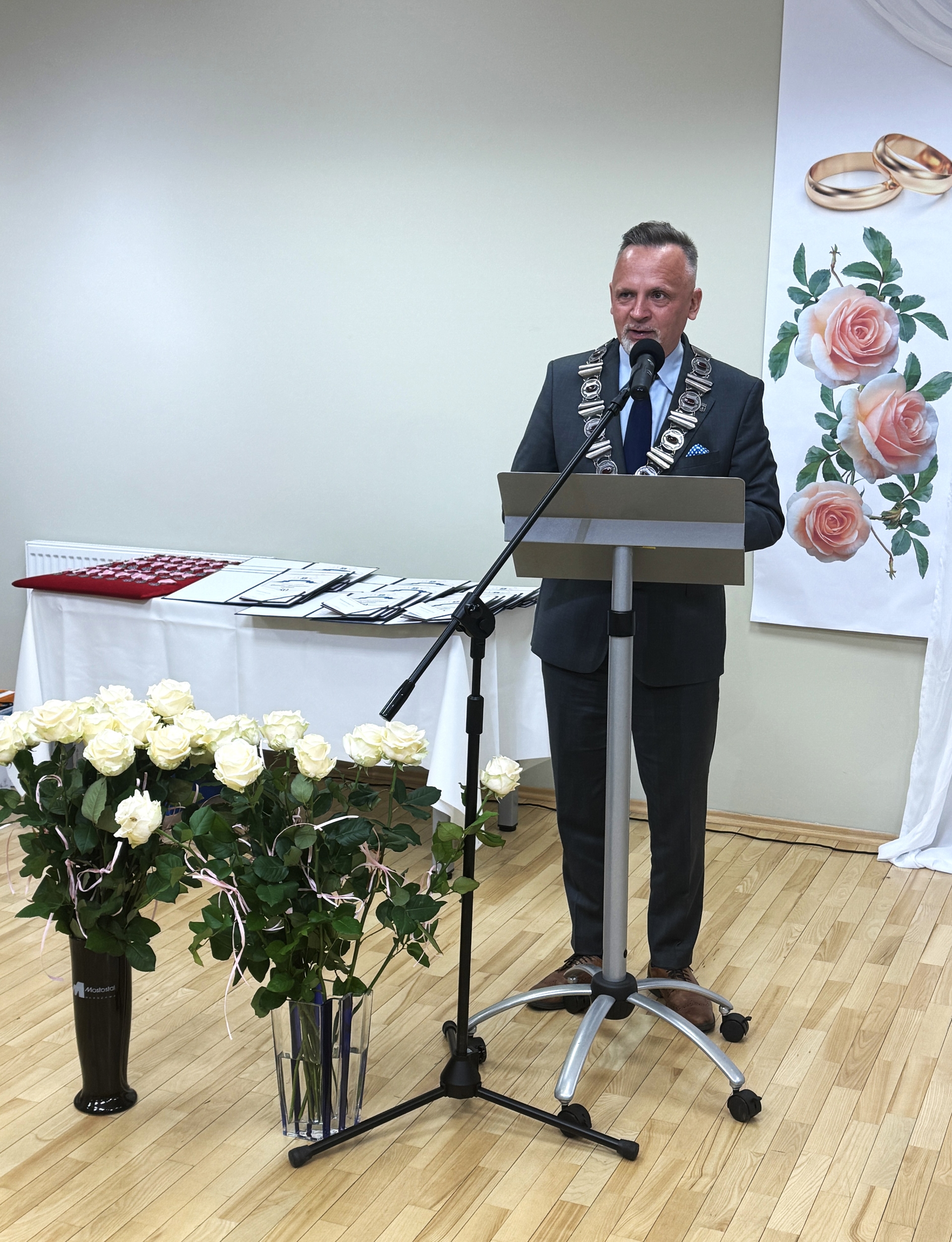 Mężczyzna w garniturze stoi za mównicą i przemawia do mikrofonu. Na jego szyi wisi łańcuch urzędowy. Przed mównicą stoją bukiety białych róż, a z tyłu widać stół z dokumentami i plakat z różami i obrączkami.