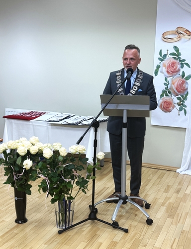 Mężczyzna w garniturze stoi za mównicą i przemawia do mikrofonu. Na jego szyi wisi łańcuch urzędowy. Przed mównicą stoją bukiety białych róż, a z tyłu widać stół z dokumentami i plakat z różami i obrączkami.