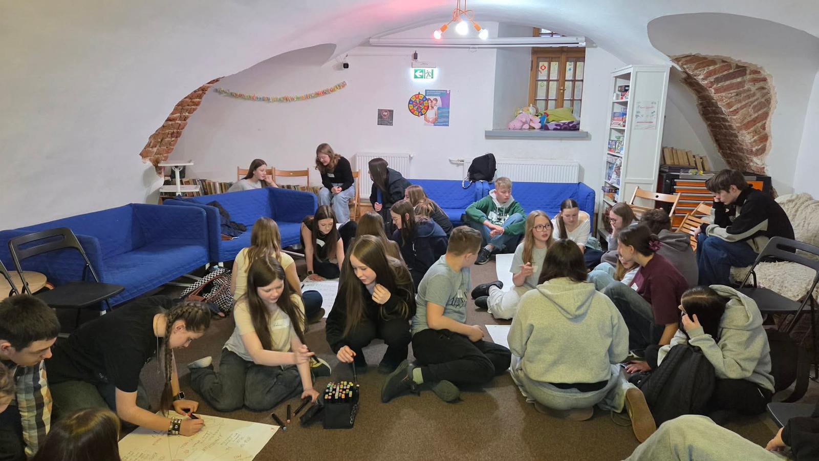 Grupa około dwudziestu nastolatków siedzi i pracuje w podziemnym pomieszczeniu. Pomieszczenie ma białe sklepione sufity z odsłoniętą czerwoną cegłą i niebieskie sofy przy ścianach. Młodzi ludzie siedzą na podłodze i krzesłach, rozkładają kartki i pisaki. Wnętrze jest oświetlone sztucznym światłem, w rogu stoi regał i okno z pluszakami.