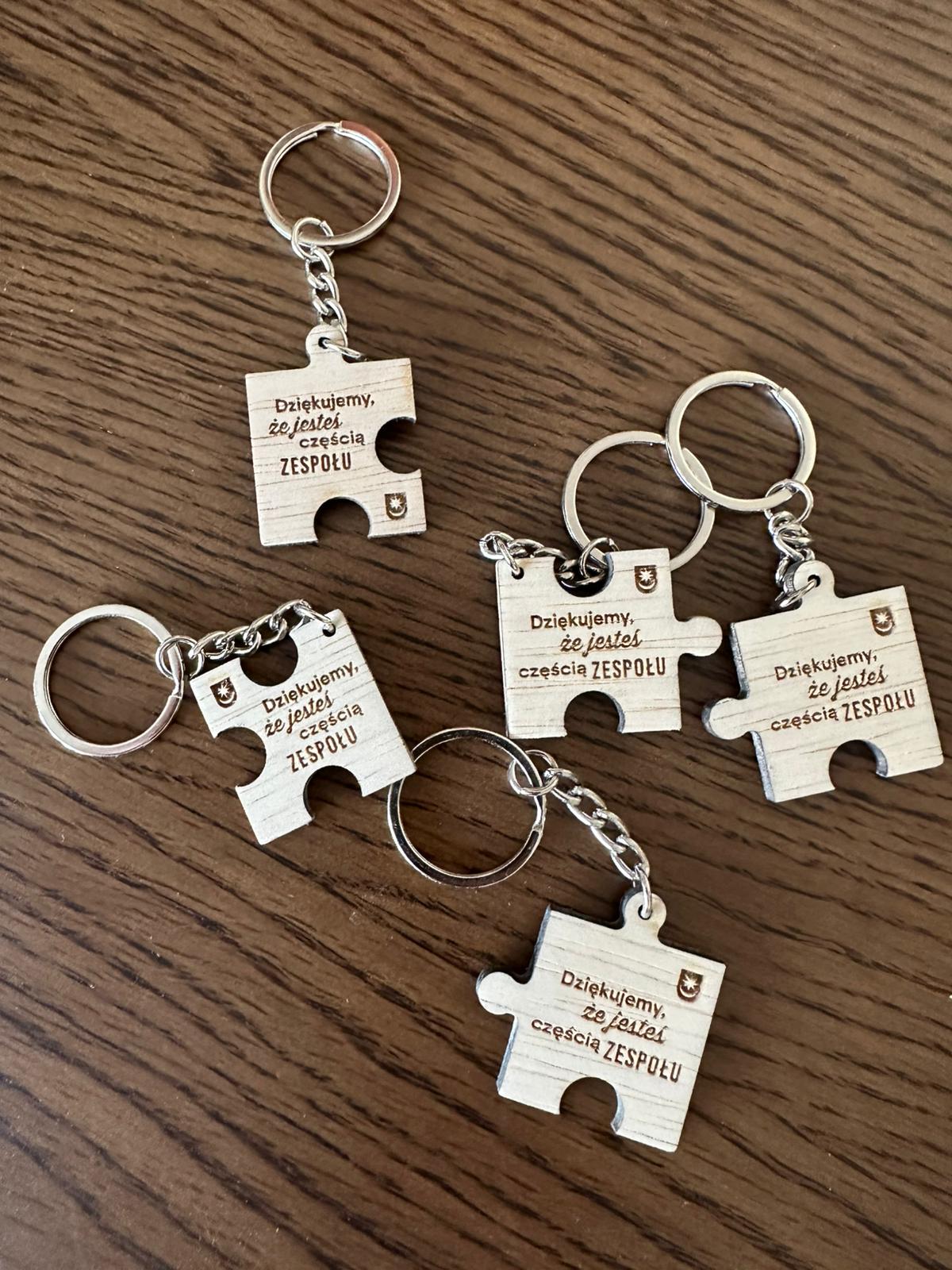 Pięć drewnianych breloków w kształcie elementów puzzli z metalowymi kółkami leży rozrzuconych na brązowym, drewnianym blacie.