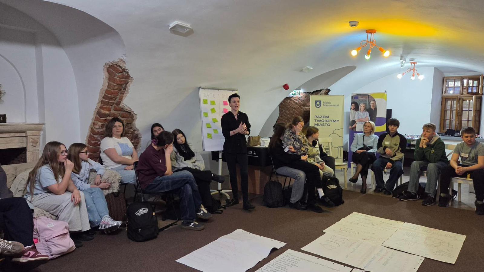 Grupa około piętnastu osób siedzi w kręgu w podziemnej sali z białym sklepionym sufitem i odsłoniętym fragmentem czerwonej cegły. Prowadzący stoi w środku przy tablicy z kolorowymi karteczkami samoprzylepnymi. Na podłodze przed kręgiem leżą duże kartki z notatkami. Z tyłu stoją dwa stojaki z banerami, a sufit jest oświetlony wielobarwnymi lampami.