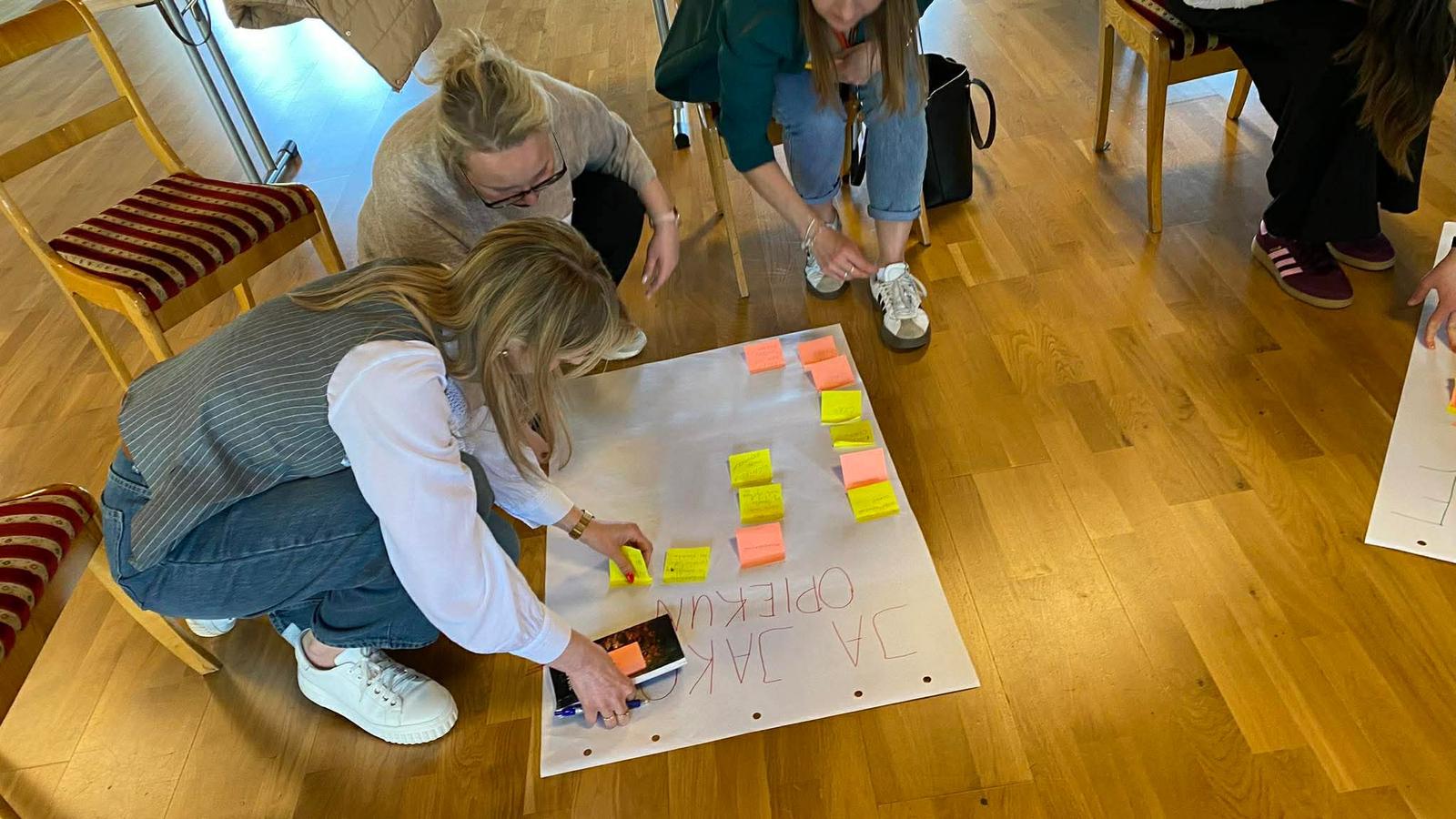 Cztery kobiety klęczą lub pochylają się nad dużą kartką leżącą na drewnianej podłodze. Przyklejają i układają żółte i różowe karteczki samoprzylepne; wokół stoją krzesła.