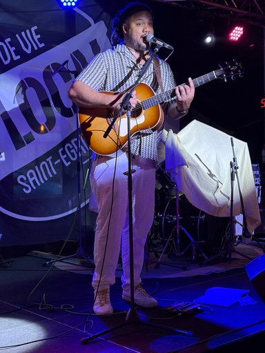 Mężczyzna śpiewa do mikrofonu i gra na akustycznej gitarze na scenie. Ma krótkie dredy, czarno-białą wzorzystą koszulę i jasne spodnie. W tle wisi baner, scenę oświetlają fioletowe i niebieskie światła.