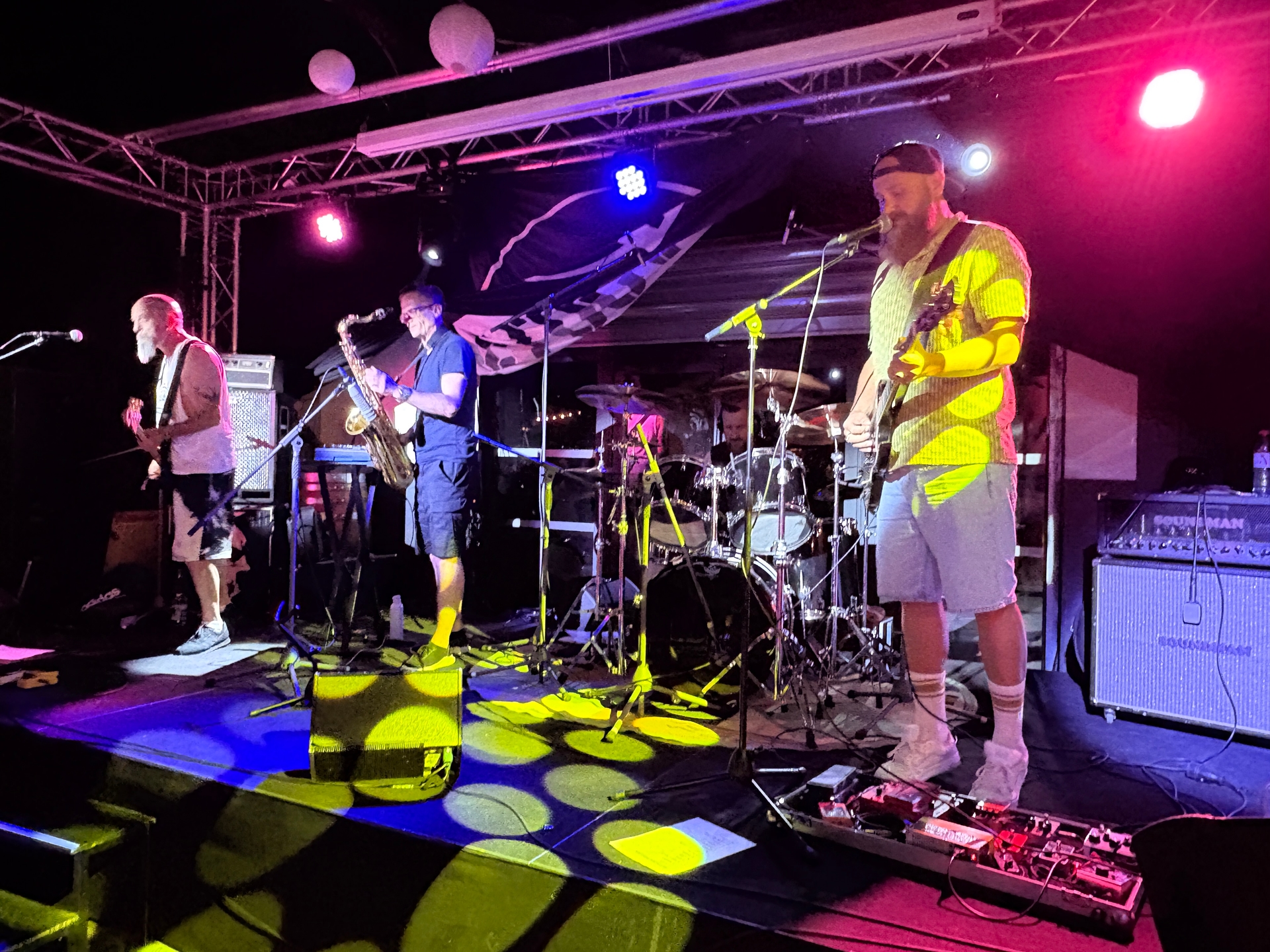 Czterech muzyków gra na oświetlonej scenie nocą. Gitarzyści stoją po bokach, saksofoniasta stoi w środku, perkusja jest z tyłu. Nad sceną widoczna metalowa konstrukcja i wiszące lampy. Tło ciemne, kolorowe reflektory żółte, różowe i niebieskie padają na podłogę.