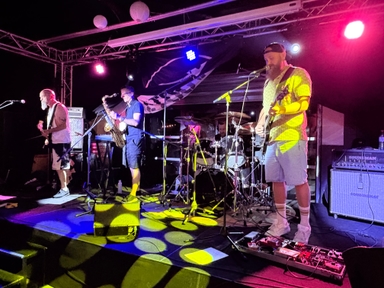 Czterech muzyków gra na oświetlonej scenie nocą. Gitarzyści stoją po bokach, saksofoniasta stoi w środku, perkusja jest z tyłu. Nad sceną widoczna metalowa konstrukcja i wiszące lampy. Tło ciemne, kolorowe reflektory żółte, różowe i niebieskie padają na podłogę.