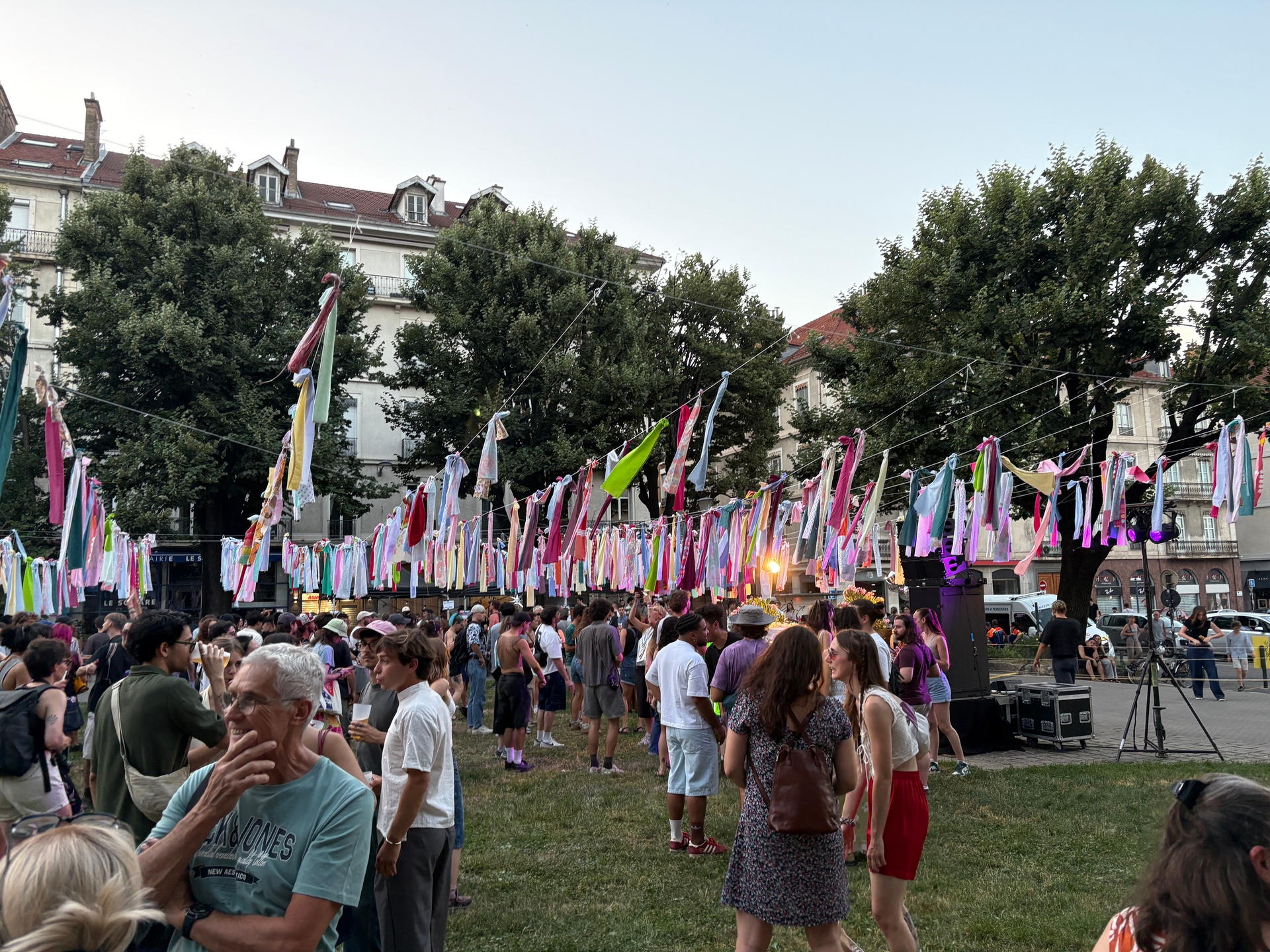 Duży tłum ludzi stoi na trawniku podczas plenerowego festiwalu. Nad głowami wiszą liczne kolorowe wstążki i pasy materiału rozpięte na sznurach, w tle wysokie drzewa i kamienice pod jasnym niebem.