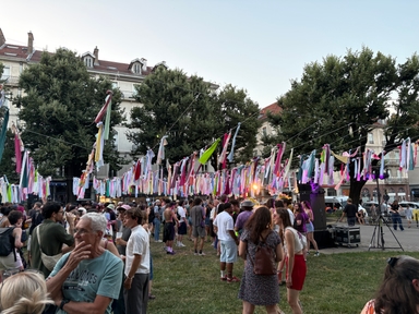 Duży tłum ludzi stoi na trawniku podczas plenerowego festiwalu. Nad głowami wiszą liczne kolorowe wstążki i pasy materiału rozpięte na sznurach, w tle wysokie drzewa i kamienice pod jasnym niebem.