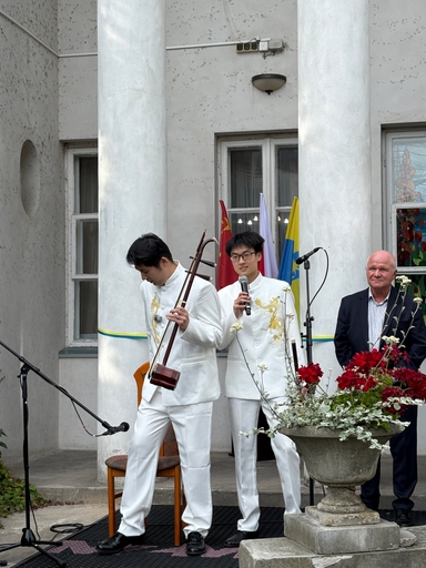 Dwóch młodych mężczyzn w białych strojach z złotym haftem stoi przed białymi kolumnami. Jeden trzyma mikrofon, drugi gra na dwustrunowym instrumencie smyczkowym. Z prawej stoi starszy mężczyzna w garniturze, w tle wiszą flagi i okna, na pierwszym planie donica z czerwonymi kwiatami.