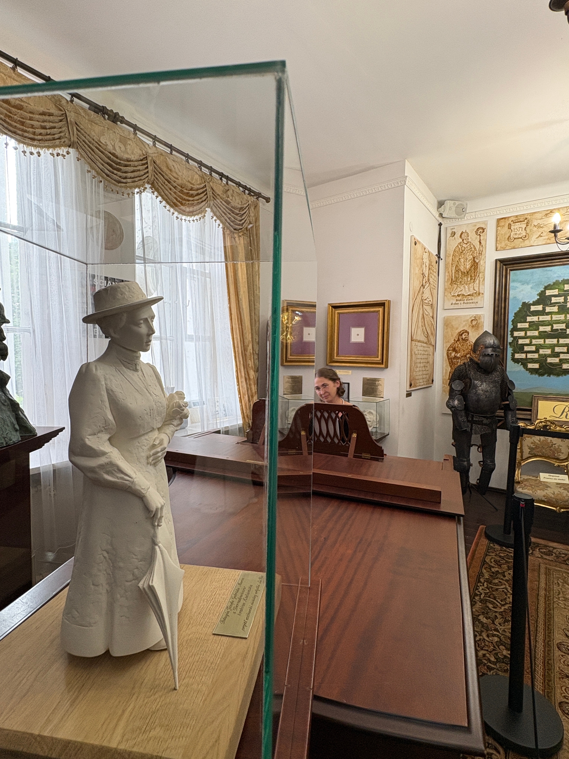 Wnętrze muzeum w dzień. Biała rzeźba kobiety w kapeluszu trzyma kwiaty i parasolkę w przeszklonej gablocie po lewej. Duży drewniany stół i oszklona przegroda biegną wzdłuż środka. Kobieta siedzi przy biurku w tle pośrodku obrazu. Po prawej stoi zbroja rycerska i ściana z oprawionymi rycinami i obrazami. Za gablotami wysokie okna z białymi firanami i złotymi zasłonami.