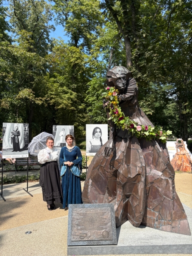 W parku stoi duża brązowa rzeźba kobiety z rozłożystą suknią i plisowanym kołnierzem ozdobiona girlandą kwiatów. Przed rzeźbą po lewej stoją dwie kobiety w historycznych strojach, jedna trzyma koronkową parasolkę. Za nimi widoczne są czarno-białe portrety na tablicach. W tle gęste zielone drzewa i słoneczne niebo.