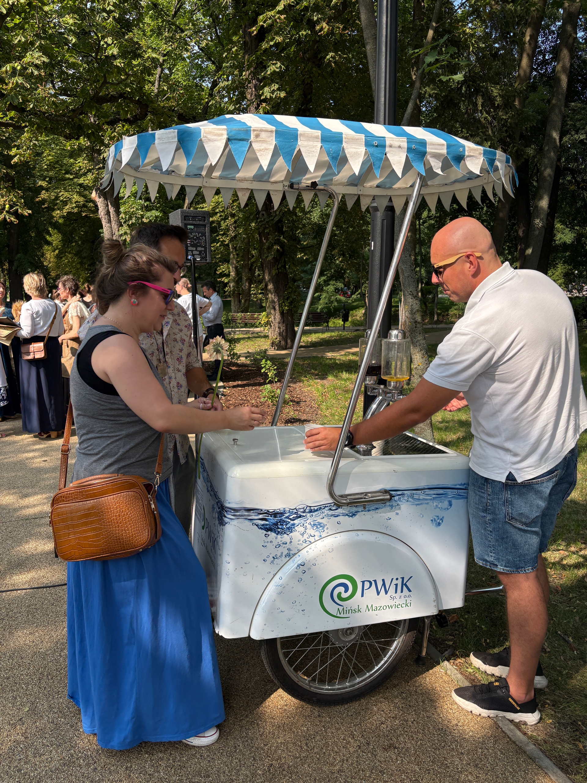 Kobieta w niebieskiej spódnicy i różowych okularach stoi przy białym wózku z niebiesko-białym parasolem. Mężczyzna w białej koszulce nalewa napój z dystrybutora, inny mężczyzna obserwuje. W tle parkowe drzewa i dzienne światło.