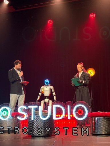 Dwie osoby stoją po bokach sceny, trzymają kartki i mikrofony. Humanoidalny robot siedzi na środku pod czerwonymi reflektorami. Przed sceną świeci duży neonowy napis, a tło jest ciemne.