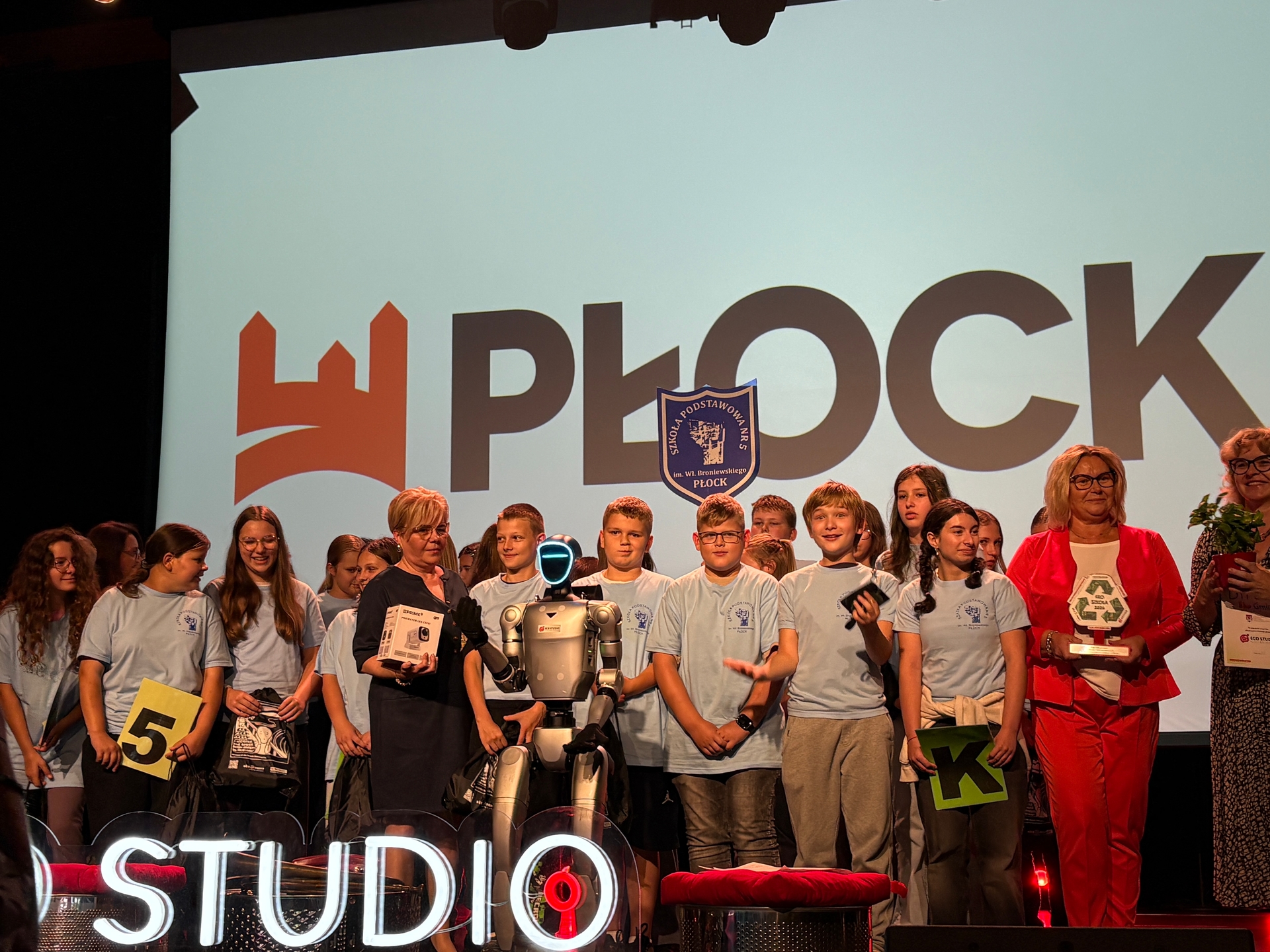Około piętnastu dzieci i kilku dorosłych stoi na scenie przed dużym białym ekranem z czarnymi literami i pomarańczowym logotypem. W centrum stoi humanoidalny robot z podświetlaną twarzą. Po prawej kobieta w czerwonym garniturze trzyma statuetkę.