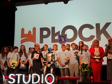 Około piętnastu dzieci i kilku dorosłych stoi na scenie przed dużym białym ekranem z czarnymi literami i pomarańczowym logotypem. W centrum stoi humanoidalny robot z podświetlaną twarzą. Po prawej kobieta w czerwonym garniturze trzyma statuetkę.