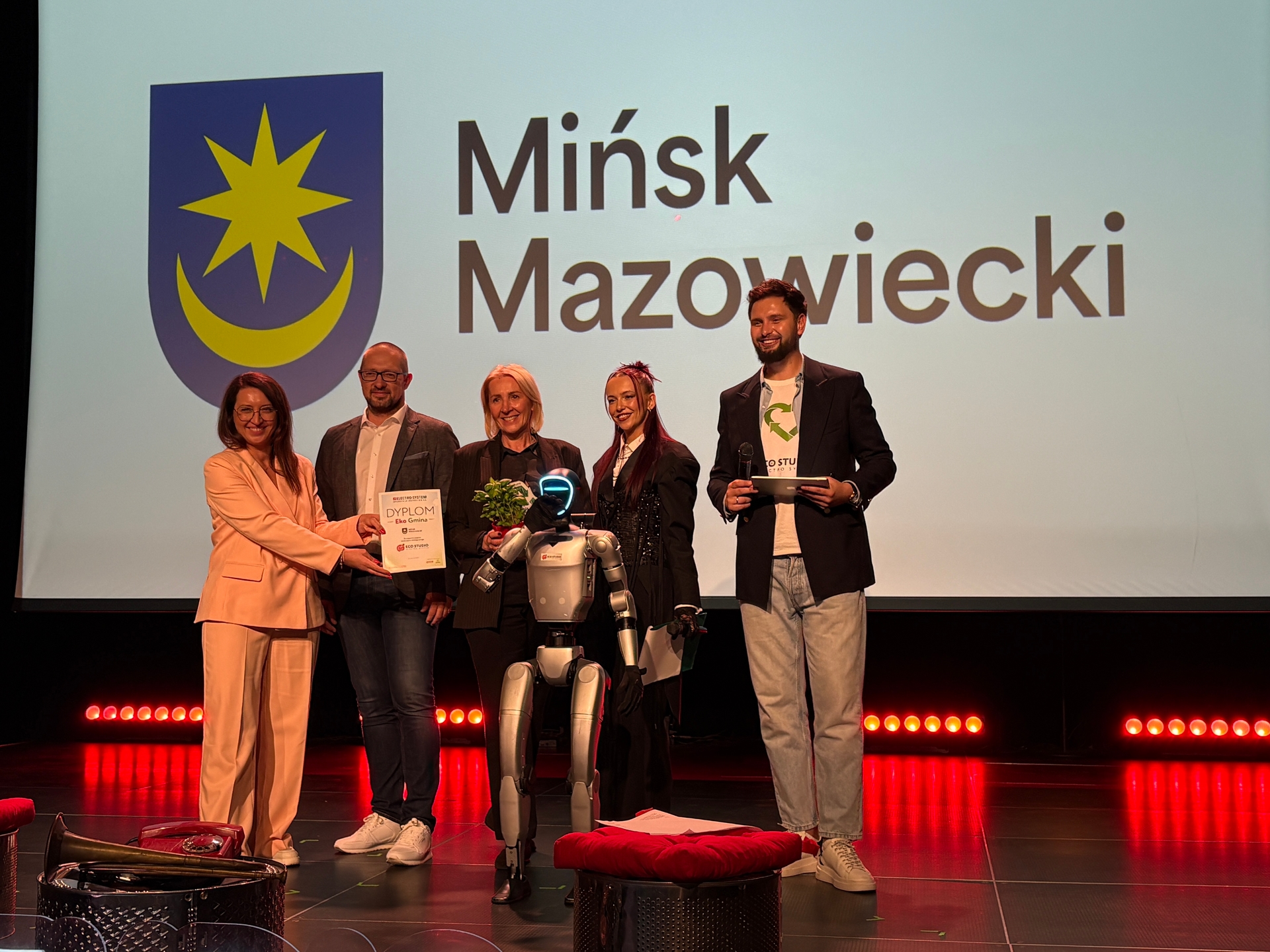 Pięć osób stoi na scenie wewnątrz przed dużym jasnym ekranem. W centrum przed grupą stoi humanoidalny robot, jedna osoba podaje dyplom, inna trzyma małą roślinę. Po lewej widoczny niebieski herb z żółtą gwiazdą i półksiężycem, po prawej duży czarny napis na białym tle. Czerwone światła oświetlają dolną część sceny.