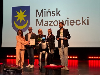 Pięć osób stoi na scenie wewnątrz przed dużym jasnym ekranem. W centrum przed grupą stoi humanoidalny robot, jedna osoba podaje dyplom, inna trzyma małą roślinę. Po lewej widoczny niebieski herb z żółtą gwiazdą i półksiężycem, po prawej duży czarny napis na białym tle. Czerwone światła oświetlają dolną część sceny.