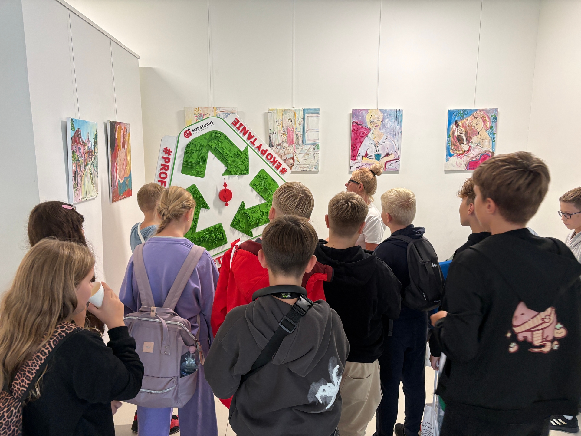 Grupa kilkunastu dzieci stoi wewnątrz jasnej galerii i ogląda obrazy zawieszone na białej ścianie. Na środku wisi duża zielona dekoracja w kształcie symbolu recyklingu. Dzieci mają plecaki i zwrócone są tyłem do aparatu.