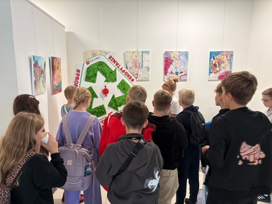 Grupa kilkunastu dzieci stoi wewnątrz jasnej galerii i ogląda obrazy zawieszone na białej ścianie. Na środku wisi duża zielona dekoracja w kształcie symbolu recyklingu. Dzieci mają plecaki i zwrócone są tyłem do aparatu.