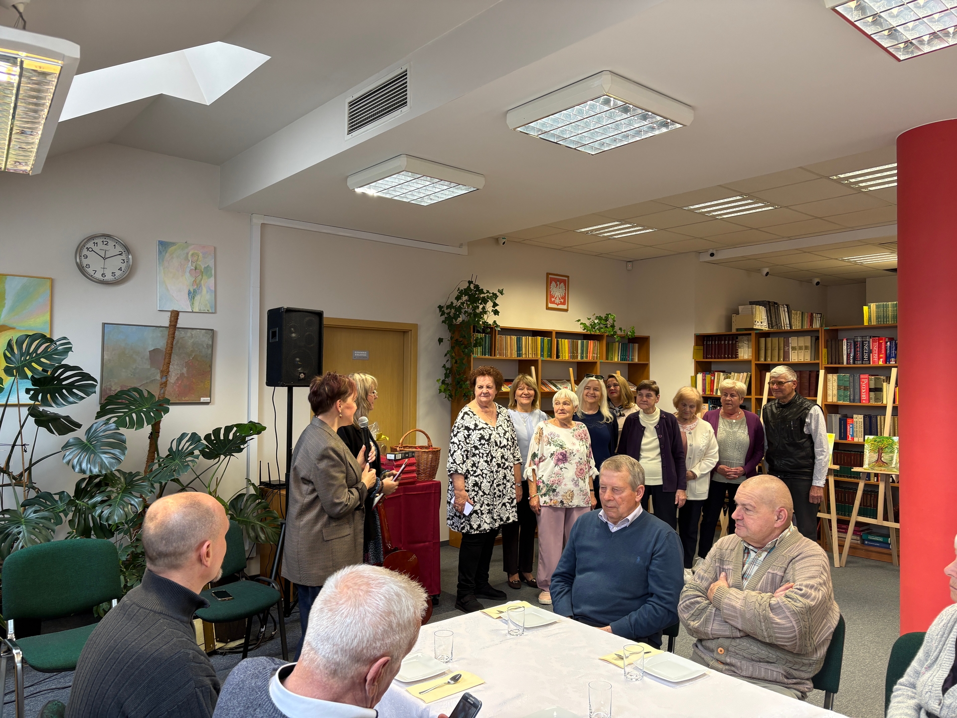 Wewnątrz biblioteki grupa około dziesięciu starszych osób stoi razem przy regałach z książkami. Z lewej kobieta mówi do mikrofonu, z przodu przy nakrytym stole siedzą trzej mężczyźni. W pomieszczeniu widać rośliny doniczkowe, zegar na ścianie i sufitowe lampy.