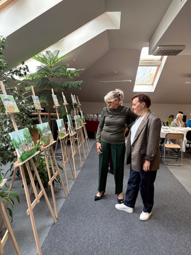 Dwie kobiety stoją w jasnym poddaszu przy rzędzie sztalug z małymi pejzażami. Nad nimi są skośne okna dachowe; po lewej rośnie wysoka zielona roślina, w tle stoi stół z filiżankami.