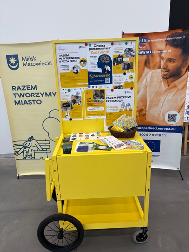 Żółty mobilny stolik z dużym czarnym kołem z lewej i małym szarym z prawej. Na górnej półce tablica z przypiętymi kolorowymi plakatami i ulotkami. Na blacie koszyk z kolorowymi materiałami i rozłożone broszury oraz gadżety. W tle stoją dwie pionowe banery: jasny baner z symbolem miasta po lewej i baner z wizerunkiem mężczyzny po prawej. Wnętrze, oświetlenie dzienne.