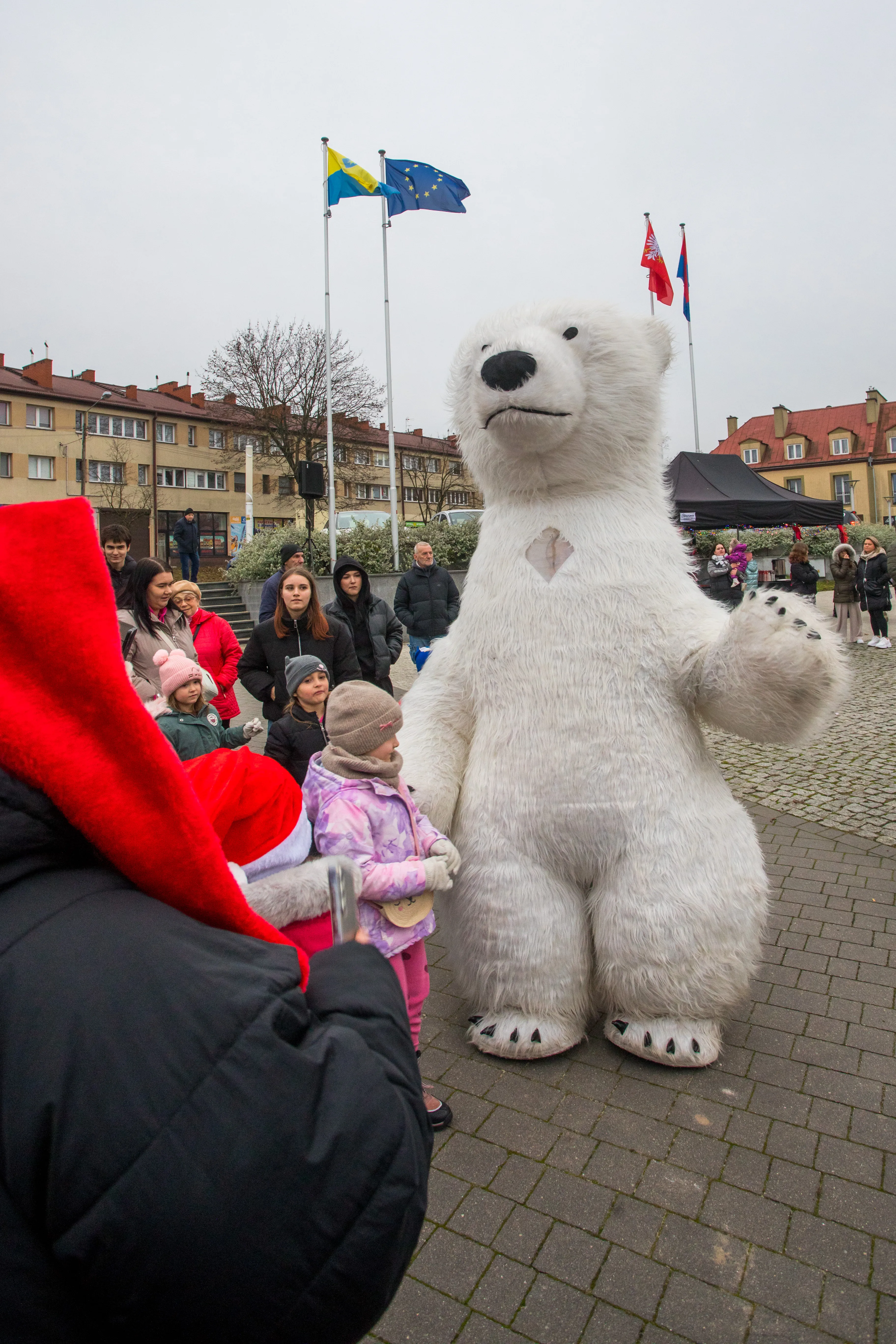 Duża postać misia polarnego stoi na brukowanym placu wśród grupy dzieci i dorosłych ubranych w zimowe ubrania. W tle widać flagi na masztach i budynki mieszkalne. Niebo jest pochmurne. Jedna osoba nosi czerwoną czapkę Mikołaja.