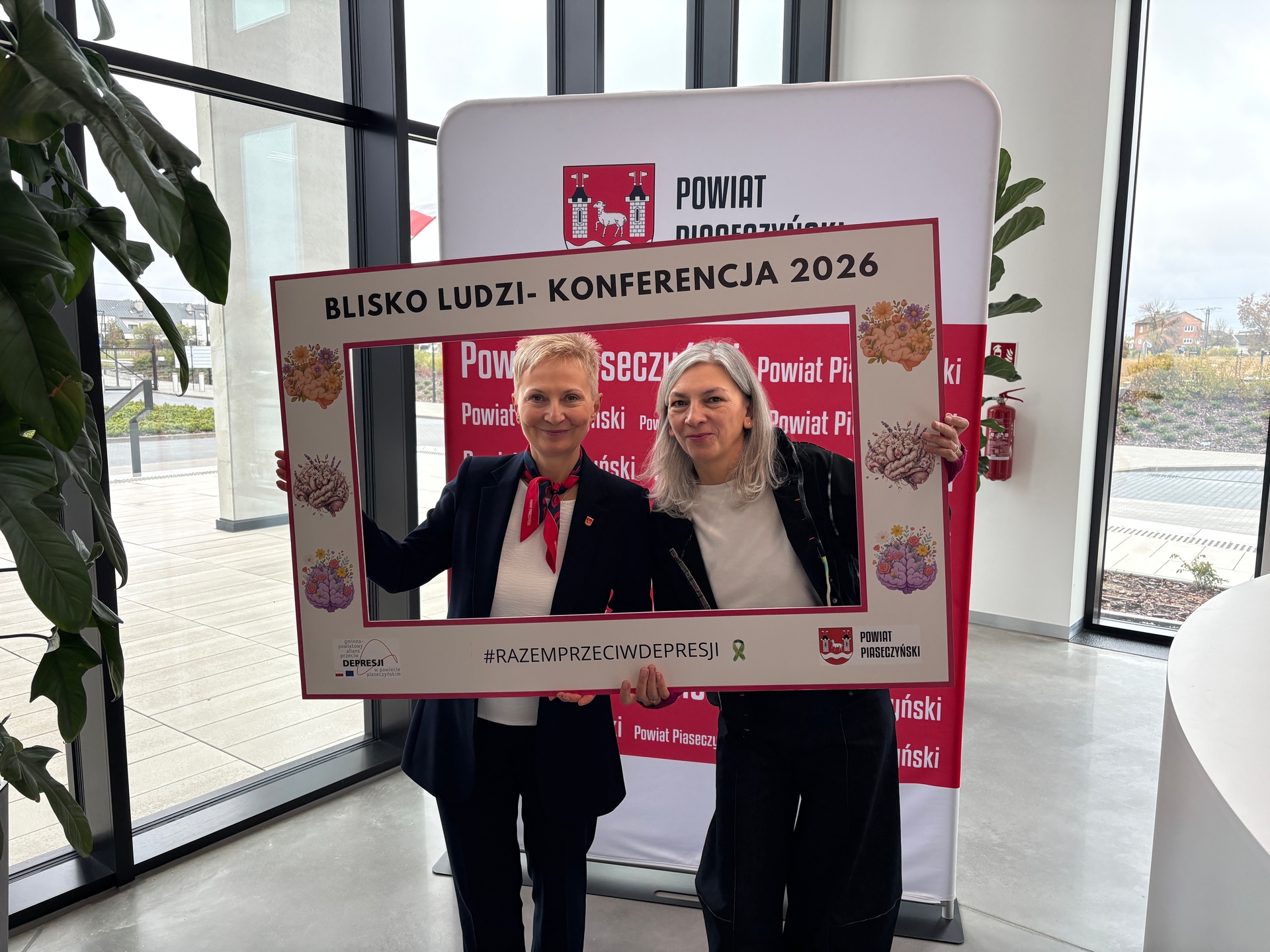 Dwie kobiety stoją blisko siebie wewnątrz przy dużych oknach i trzymają ozdobną kartonową ramę promującą wydarzenie. W tle pionowy baner z herbem, rośliny i widok na zewnątrz przez szyby, dzień.