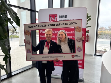 Dwie kobiety stoją blisko siebie wewnątrz przy dużych oknach i trzymają ozdobną kartonową ramę promującą wydarzenie. W tle pionowy baner z herbem, rośliny i widok na zewnątrz przez szyby, dzień.