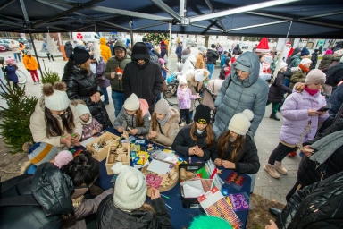 Grupa dzieci i dorosłych siedzi i stoi przy stole pod namiotem na zewnątrz zimą. Dzieci wykonują prace plastyczne, mają czapki i kurtki zimowe. W tle tłum ludzi, choinka i dekoracje świąteczne.
